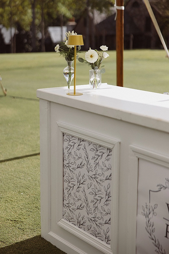 Wedding bar setup with a white portable wedding bar topped with clear bud vases of white flowers and greenery on a lawn under tent poles near trees
