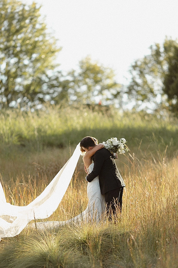 Wedding kiss portrait of bride and groom kissing in a tall grass field, her long veil blowing as she holds a white bouquet at sunset