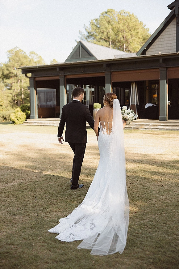 Bride and groom walking hand in hand, wedding dress with long veil and bouquet, groom in black tuxedo on an estate lawn by a modern house