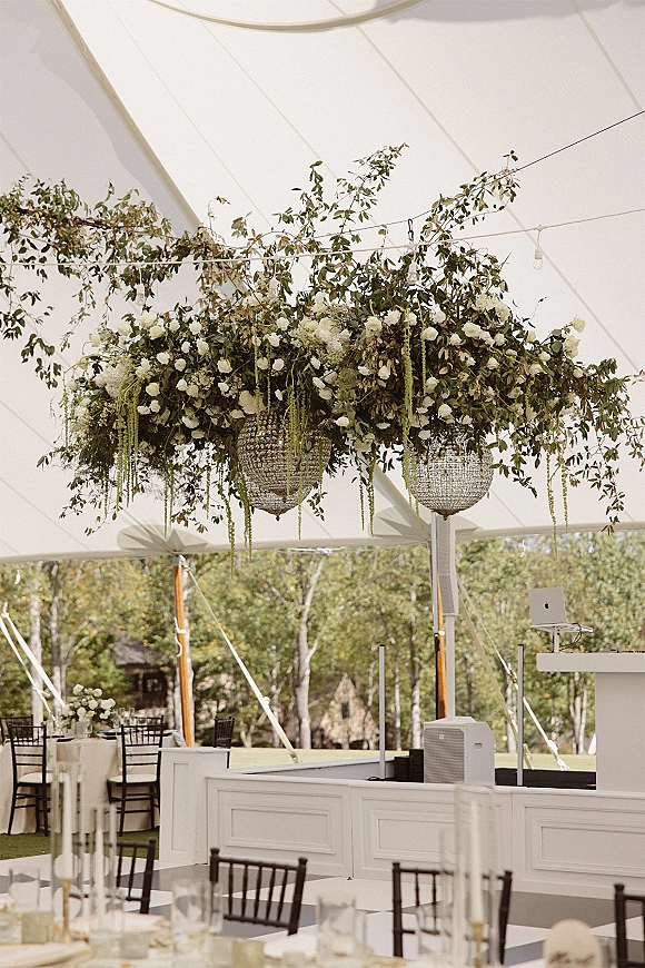 Reception decor with hanging floral installation of greenery and white blooms beneath crystal chandeliers in a draped white tent with string lights