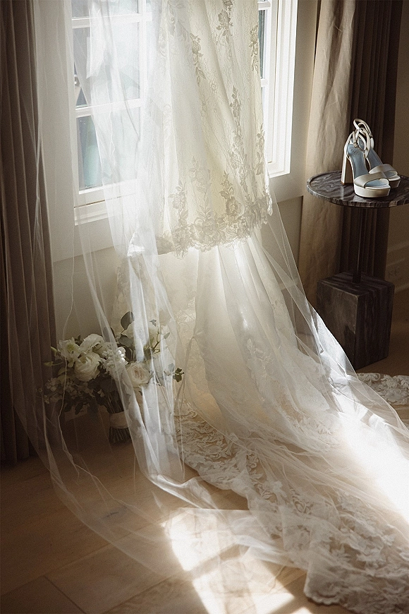 Wedding dress lace wedding gown with long train and veil, paired with white rose bouquet and heels by a window with sheer curtains