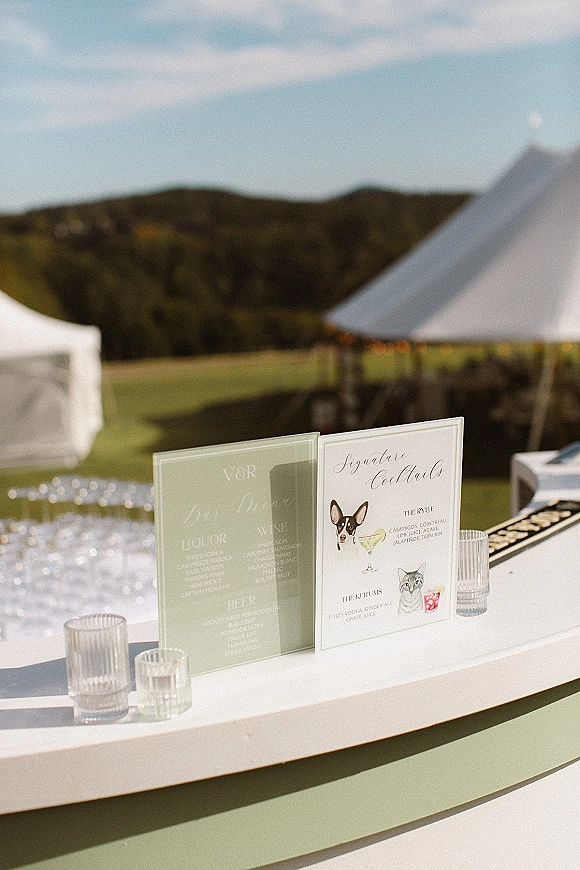 Wedding bar signage with a signature cocktail sign in acrylic holders, custom drink illustration and glass votive candles on an outdoor lawn bar under a white tent