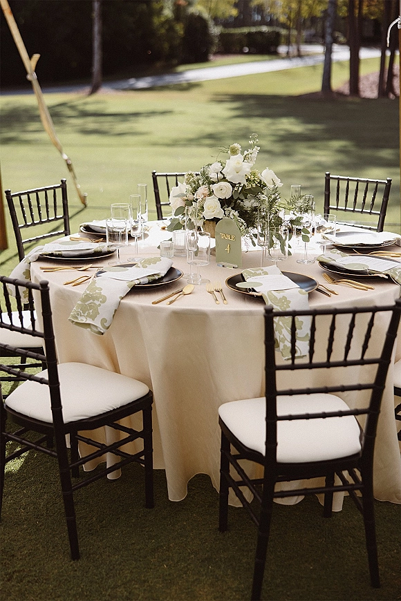 Reception tablescape with an outdoor wedding reception table, ivory linens, white roses and greenery centerpiece, black chargers and gold flatware on a sunny lawn
