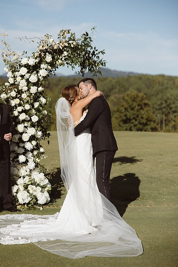 Wedding kiss beneath a floral arch with white roses and greenery, bride in lace dress and veil with train on a sunny lawn backdrop