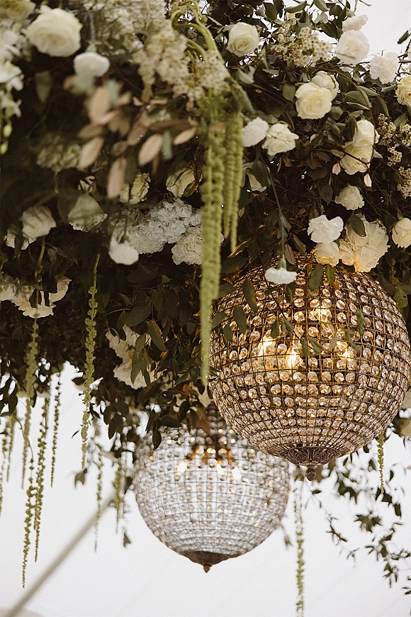 Hanging floral chandelier with wedding chandelier decor, white roses, hydrangeas, greenery and crystals glowing under a white tent ceiling