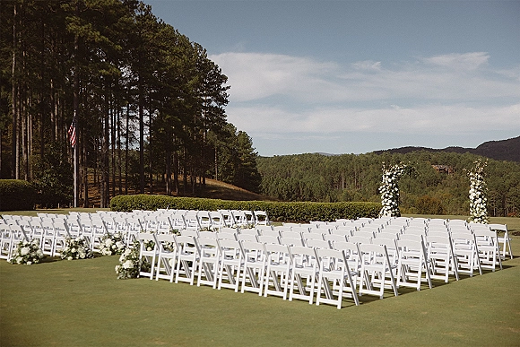 Outdoor ceremony setup with white folding chairs lining a floral aisle, leading to a white flower arch on a lawn with pine trees and hills beyond