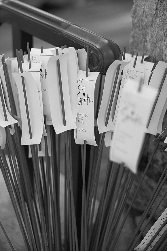 Wedding sparkler tags with handwritten calligraphy clipped to sparklers, arranged on a basket handle against a tree trunk backdrop