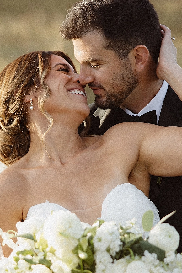 Wedding couple portrait of bride and groom nose to nose, her strapless lace gown and white bouquet glowing in golden hour outdoors