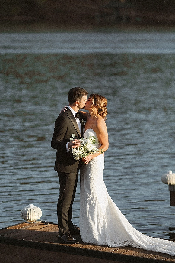 Wedding kiss as bride and groom embrace on a wooden dock by the lake, her strapless lace dress and bouquet framed by dock lights