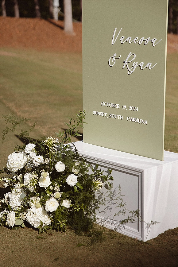 Wedding welcome sign in clear acrylic with raised lettering, white roses and hydrangeas with greenery on a pedestal in a sunny lawn field