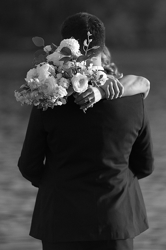 Wedding couple portrait of bride and groom embrace from behind, bouquet of white roses and greenery over her shoulder in outdoor landscape