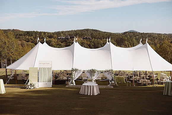 Wedding reception tent with sailcloth wedding tent string lights, round tables, bar, and greenery garlands on a lawn with hills and blue sky