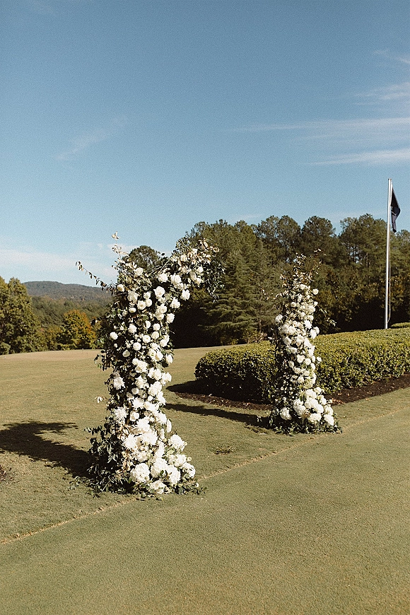 Wedding ceremony arch with white flowers and greenery on floral pillars, set on a manicured lawn with hills and blue sky beyond