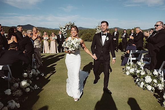 Ceremony recessional with bride and groom walking aisle, holding hands with a white rose bouquet, guests cheering on a mountain lawn backdrop