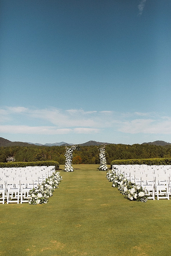 Outdoor ceremony setup with wedding aisle flowers lining a straight aisle of white chairs, framed by floral pillars on a lawn with mountain views