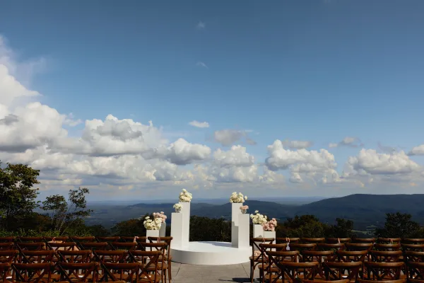 Ceremony setup for outdoor wedding ceremony with white pedestal plinth florals and circular platform, cross-back chairs on a mountain-view patio
