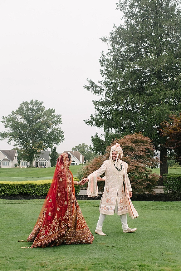 Couple portrait of an Indian wedding couple holding hands on a green lawn, bride in red lehenga with embroidered dupatta veil under overcast sky