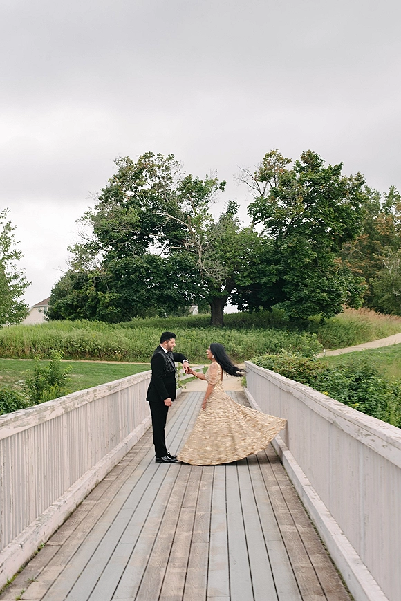 Couple portrait of bride twirling dress, holding hands with groom in black suit on a wooden bridge with white railings under overcast sky