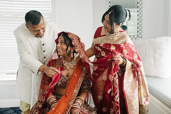 Bridal getting ready as bride in red lehenga sits by window light while mother adjusts dupatta, showcasing mehndi hands and jewelry.