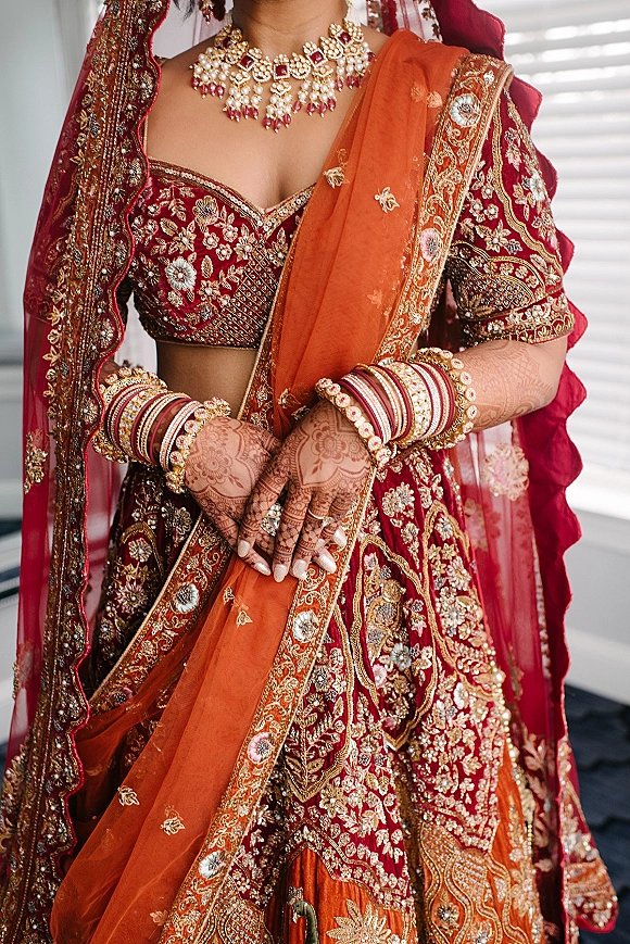 Bridal lehenga and red bridal lehenga with heavy embroidery, orange dupatta, and gold jewelry, shown indoors by window blinds