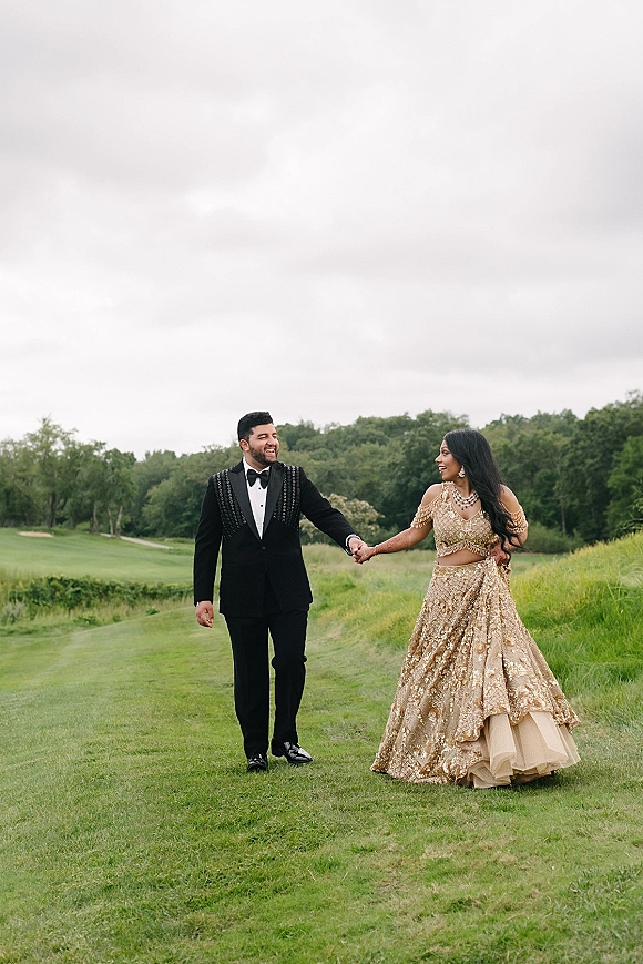 Couple portrait of bride and groom holding hands, her gold lehenga and his black tuxedo, walking in a green field under cloudy sky