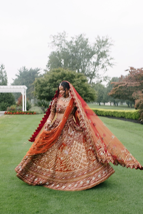 Bridal portrait of a South Asian bride in a red lehenga with gold embroidery, twirling on a garden lawn near a white pergola