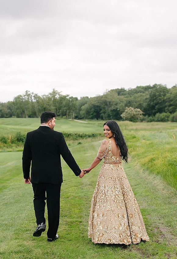 Couple portrait of bride and groom holding hands walking away as she looks back, her gold embellished lehenga in a grassy field under cloudy sky