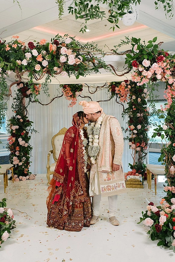 Wedding kiss between a south asian wedding couple under a rose and greenery arch, bride in embroidered red lehenga with veil, indoor stage backdrop