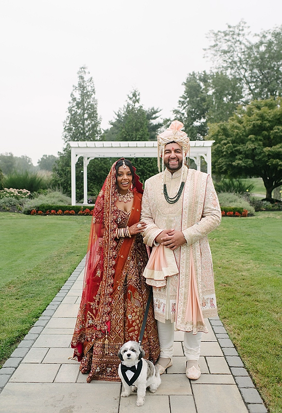 Couple portrait of a south asian wedding couple holding hands with their tuxedo dog on a garden lawn by a white pergola