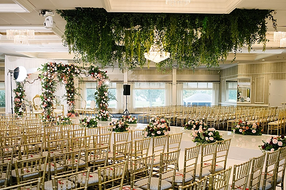 Ceremony setup with a floral arch of pink and white roses, gold Chiavari chairs, and chandelier under hanging greenery in a ballroom