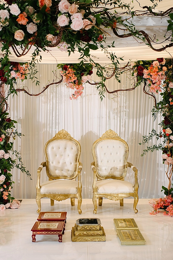 Wedding ceremony backdrop with a floral arch of roses and greenery, hanging blooms and branches over gold and white chairs on an indoor stage