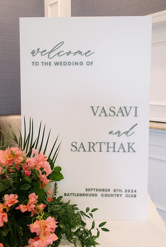 Wedding welcome sign with calligraphy lettering and a pink floral arrangement with palm fronds, set indoors against wall paneling