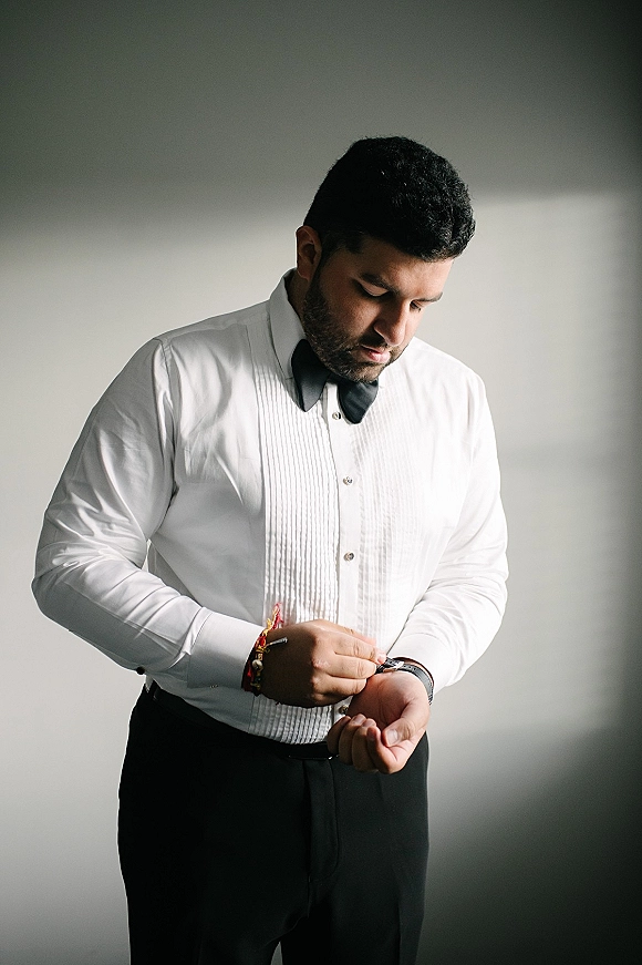 Groom portrait in a white tuxedo shirt with black bow tie, checking his wristwatch and bracelets in soft indoor natural light