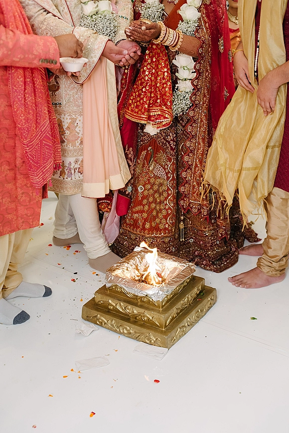 Wedding ceremony ritual with bride in red lehenga and groom holding hands beside a sacred fire altar, garlands and petals on white floor
