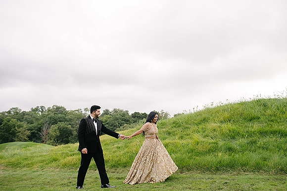Couple portrait of bride and groom holding hands, walking on a grassy hill as her embellished gold lehenga and his tuxedo stand out under overcast sky