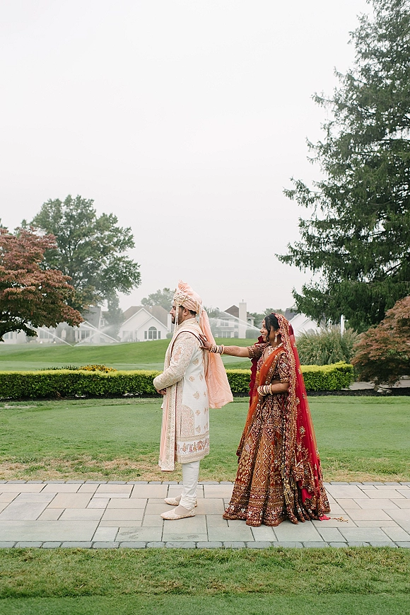 First look moment as a bride in a red lehenga taps the groom’s shoulder on a stone walkway, her dupatta veil draped behind on a lawn