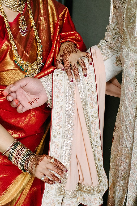 Wedding hands close-up with bridal mehndi hands, gold rings and bangles resting on groom’s embroidered sherwani sleeve against a dark backdrop