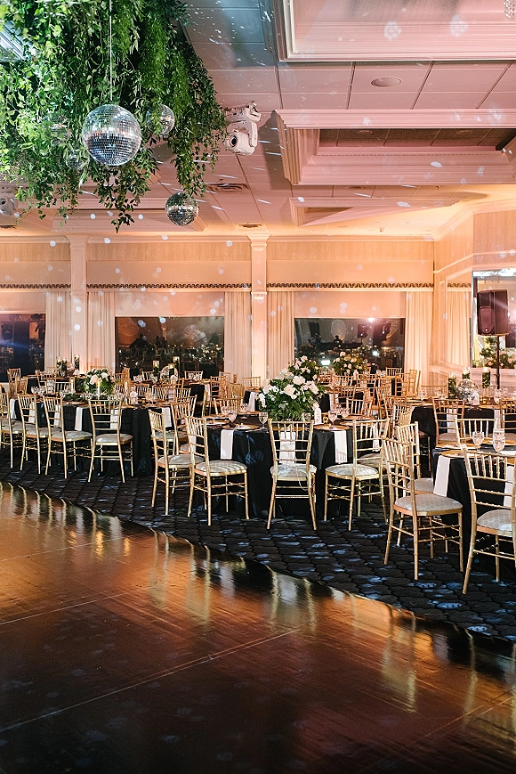 Reception tablescape with black and gold wedding reception decor, tall white floral centerpieces, votive candles, and disco balls in a ballroom at night
