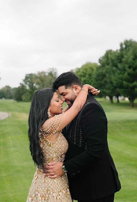 Couple portrait of bride and groom embracing with a forehead touch, her gold embellished dress and mehndi hands on a green lawn under overcast sky