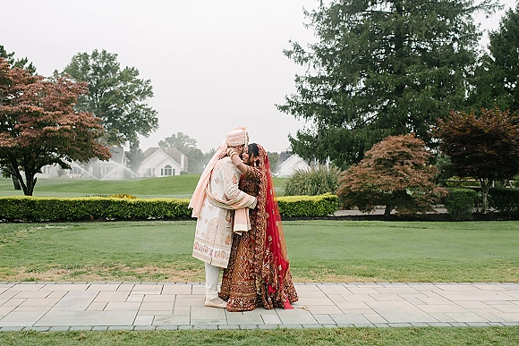 Couple portrait of a South Asian wedding couple embracing, bride in red lehenga and dupatta veil, on a golf course lawn with sprinklers