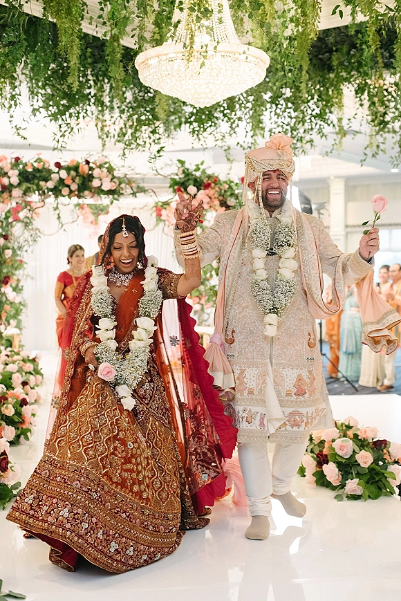 Wedding recessional as Hindu newlyweds walk hand in hand in red lehenga and sherwani, floral garlands under a chandeliered mandap.