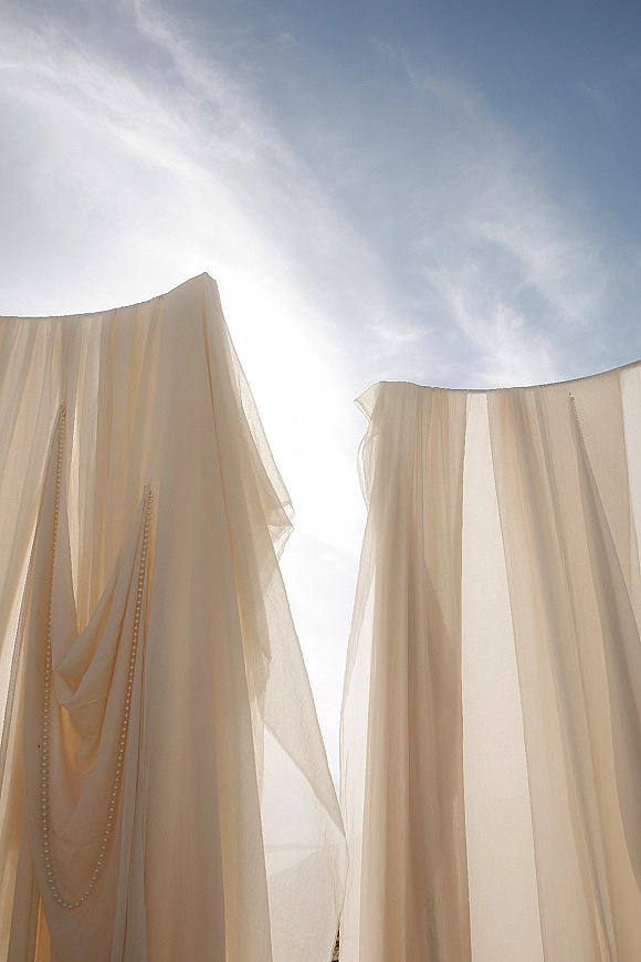 Wedding dress detail with button back wedding dress design, sheer tulle train and beaded trim backlit by sunlight against blue sky
