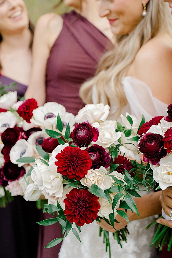 Bridal bouquet of burgundy and white bouquet with dahlias and ranunculus, held before bridesmaids in burgundy dresses and blurred greenery.
