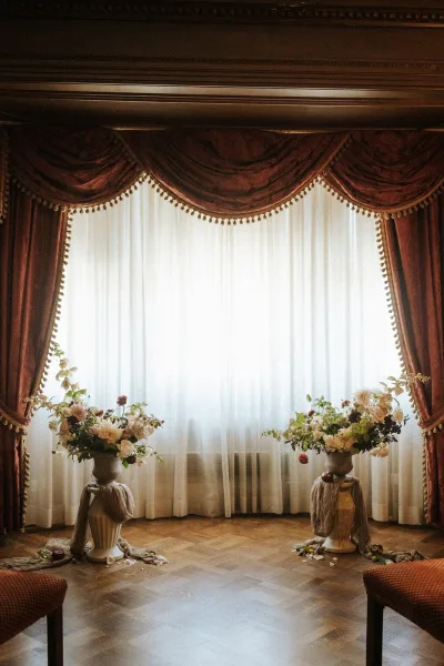 Ceremony backdrop styled for indoor ceremony decor, anchored by classic urn vases and framed by sheer curtains and a large window.