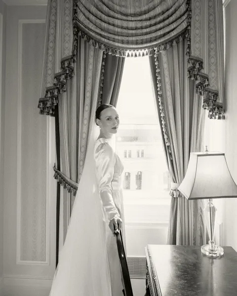 Bridal portrait of a bride by window, gazing toward city buildings as a sheer veil catches the light beside drapery and a paneled wall.