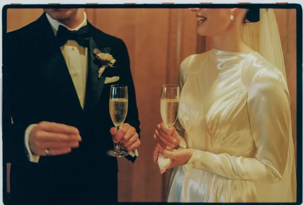 Wedding champagne toast as the bride and groom cheer, her satin bridal gown gleaming against a warm wood-paneled wall.