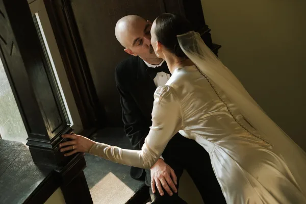 Wedding kiss portrait as a bride and groom kiss beside a wood-trimmed window, her sheer veil glowing in soft natural light indoors.
