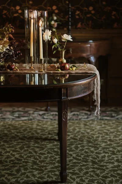 Wedding centerpiece with a taper candle centerpiece in brass candlesticks, arranged on a dark wood table against moody floral wallpaper.