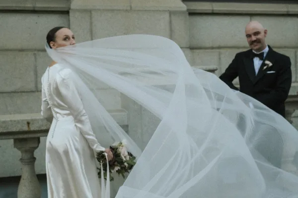 Bridal portrait of a bride with long veil sweeping behind her, posed by a stone balustrade and stone wall.