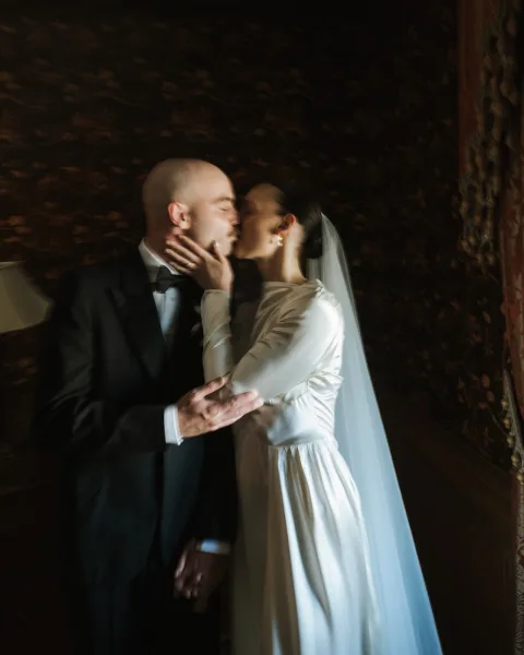Wedding kiss portrait as a bride and groom kiss, holding hands, her veil brushing his cheek as patterned wallpaper and drapery glow behind them.